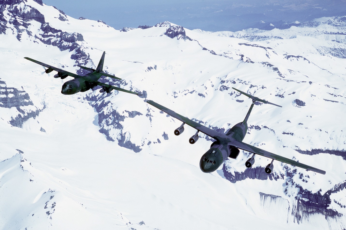 An air-to-air front view of a C-141 Starlifter aircraft (right) from the 62nd Military Airlift Squadron, and a C-130 Hercules aircraft above snow covered mountains - U.S. National Archives & DVIDS Public